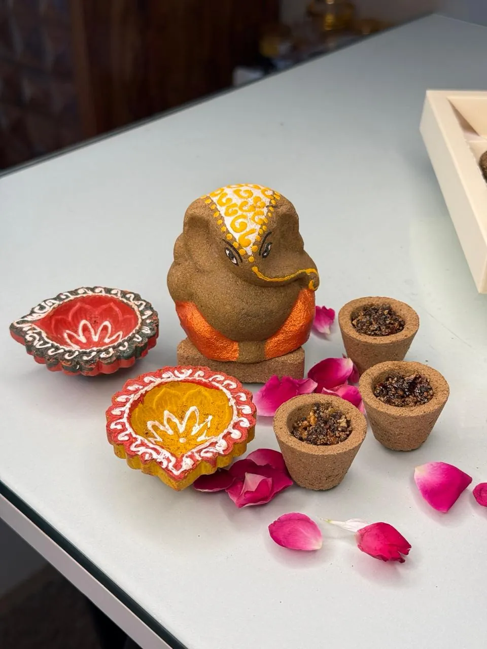 Hand-painted clay Ganesha, two painted diyas and three small cups of incense on a table scattered with rose petals