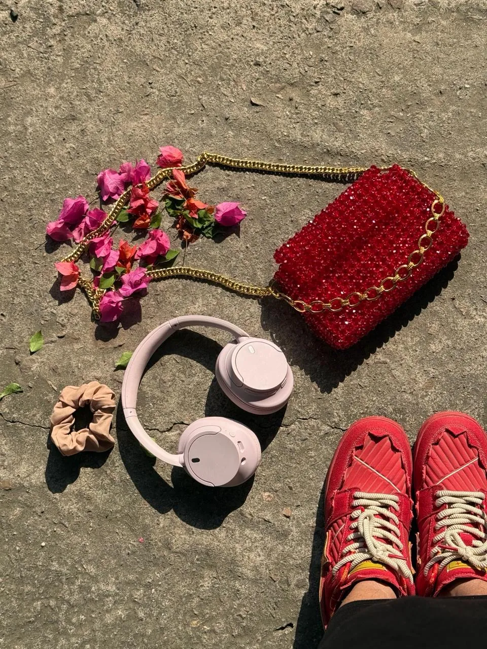 Flat lay of a red beaded chain-strap bag, pink headphones, a scrunchie and bougainvillea petals beside red sneakers