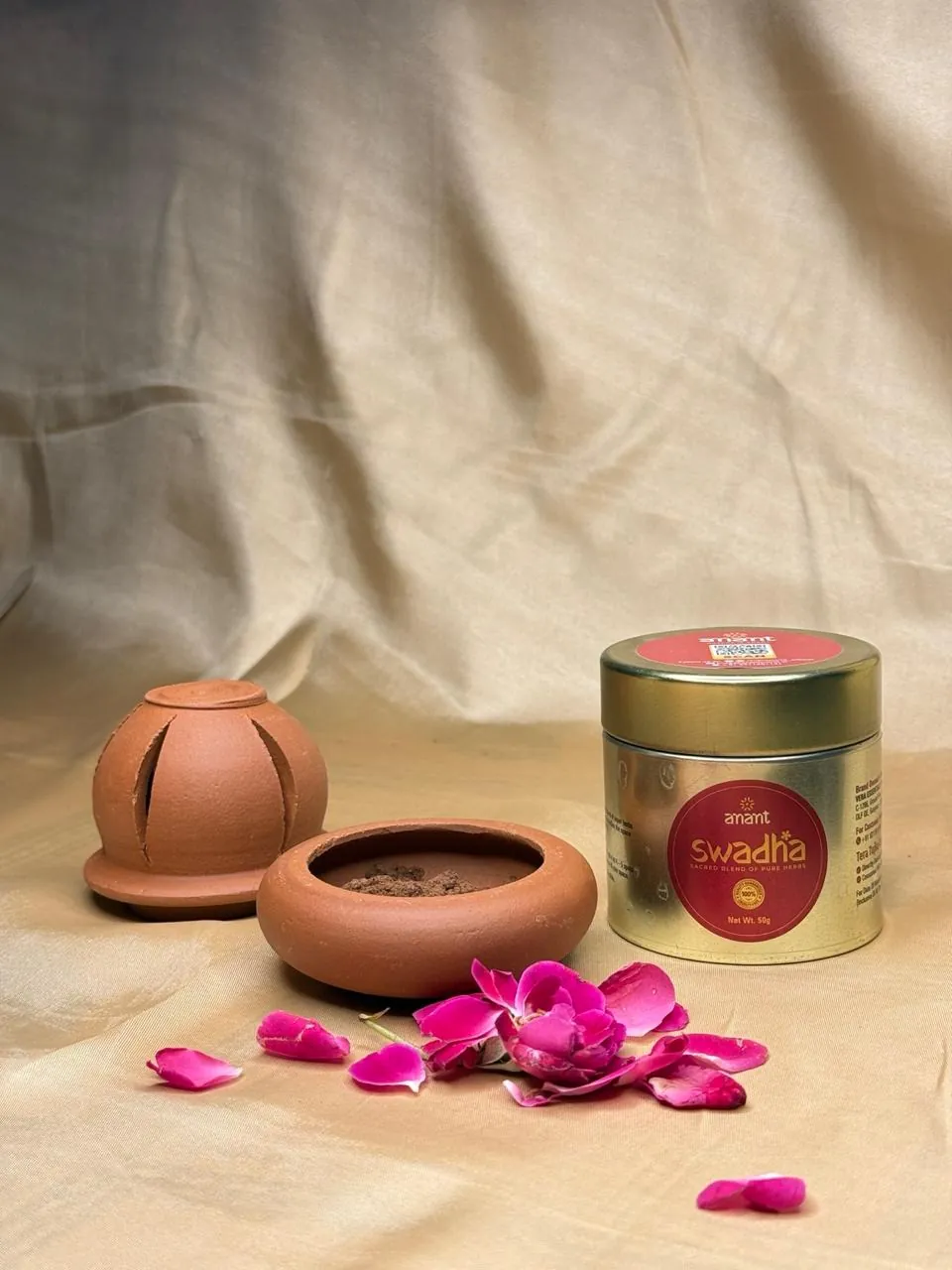 Swadha herbal blend tin by Anant World beside a terracotta lantern-style burner and an open clay bowl, with rose petals on gold silk